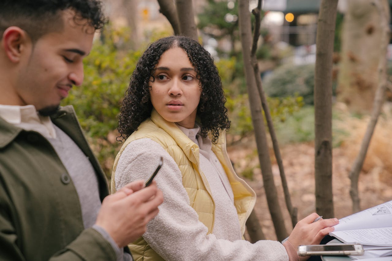 latin couple outside and the girl is looking at the boyfriend who is smiling at his phone, she is displaying being jealous